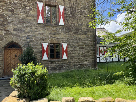Burghaus Scheltensülz Altes, steinernes Haus mit rot-weißen Fensterläden, umgeben von grünem Rasen und Bäumen.