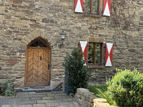 Burghaus Scheltensülz Eingang Altes Steinhaus mit Holztür und roten Fensterläden in malerischer, grüner Umgebung.