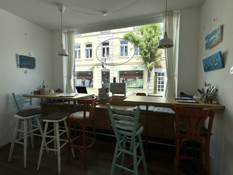Kaffeepause - Innenansicht Gemütliches Atelier mit hoher Fensterfront, Blick auf Straße, bestückt mit Holzstühlen und Malutensilien.