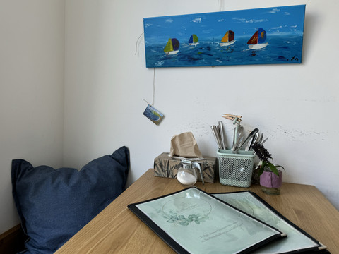 Kaffeepause - Innenansicht Ein Holztisch mit Speisekarten, daneben ein Kissen und ein buntes Bootsgemälde an der Wand.