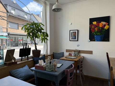 Kaffeepause - Innenansicht Gemütliches Café mit Holzmöbeln, Kunst an weißen Wänden und Topfpflanze am Fenster.