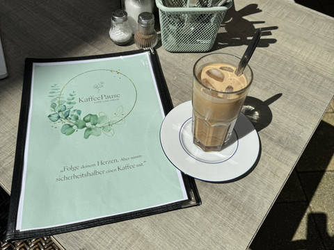 Kaffeepause - Ansicht Eiskaffee Ein Glas Eiskaffee mit Löffel steht auf einem Tisch neben einer Speisekarte des Cafés "KaffeePause".