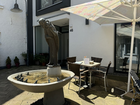 Kaffeepause - Ansicht Außenterrasse Brunnen mit zwei Figuren, umgeben von Tauben, neben Café mit Schirmen und Sitzgelegenheiten.