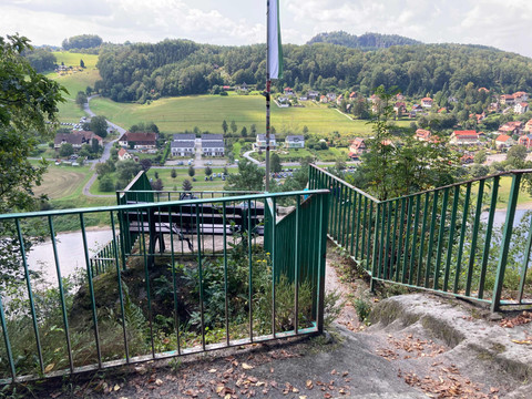 "Kleine_Bastei"_uitzichtspunt Grünes Geländer führt zu einer Aussichtsplattform mit Bank, im Hintergrund eine ländliche Landschaft mit Häusern und Hügeln unter bewölktem Himmel.