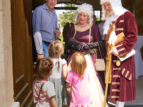Das Bild zeigt zwei Erwachsene in barocken Kostümen mit einigen Kindern im Eingangsportal des Jagdhauses.The picture shows two adults in baroque costumes with some children in the entrance portal of the hunting lodge.Na obrázku jsou dva dospělí v barokních kostýmech s několika dětmi u vstupního portálu loveckého zámečku.Zdjęcie przedstawia dwoje dorosłych w barokowych strojach z dziećmi w portalu wejściowym do domku myśliwskiego.De foto toont twee volwassenen in barokke kostuums met enkele kinderen in het toegangsportaal van het jachthuis.L'immagine mostra due adulti in costume barocco con alcuni bambini nel portale d'ingresso della casa di caccia.