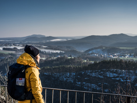 Person in gelber Jacke betrachtet von Aussichtspunkt verschneite Landschaft und Wald im Winter.