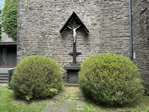 Kirche St. Walburga Steinmauer einer Kirche mit einem hölzernen Kruzifix, umgeben von zwei symmetrischen Büschen.