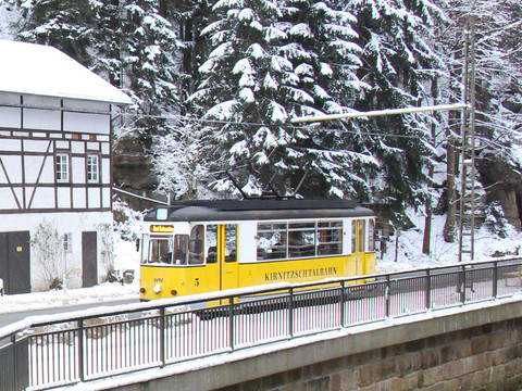Eine gelbe Straßenbahn der Kirnitzschtalbahn fährt durch eine verschneite Waldlandschaft am Beuthenfall.A yellow Kirnitzschtalbahn streetcar travels through a snow-covered forest landscape at the Beuthen Falls.Žlutá tramvaj Kirnitzschtalbahn projíždí zasněženou lesní krajinou u Beuthenských vodopádů.Żółty tramwaj Kirnitzschtalbahn przejeżdża przez pokryty śniegiem leśny krajobraz przy wodospadzie Beuthen.Een gele Kirnitzschtalbahn tram rijdt door een besneeuwd boslandschap bij de Beuthen watervallen.Un tram giallo della Kirnitzschtalbahn attraversa un paesaggio forestale innevato presso le cascate di Beuthen.