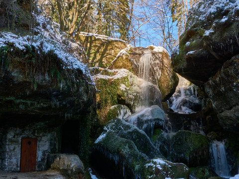 Verschneiter Wasserfall im Wald mit moosbewachsenen Steinen und einem kleinen, versteckten Eingang links.Snow-covered waterfall in the forest with moss-covered stones and a small, hidden entrance on the left.Zasněžený vodopád v lese s mechem porostlými kameny a malým skrytým vchodem vlevo.Pokryty śniegiem wodospad w lesie z pokrytymi mchem kamieniami i małym, ukrytym wejściem po lewej stronie.Besneeuwde waterval in het bos met met mos bedekte stenen en een kleine, verborgen ingang aan de linkerkant.Cascata innevata nel bosco con pietre ricoperte di muschio e un piccolo ingresso nascosto sulla sinistra.