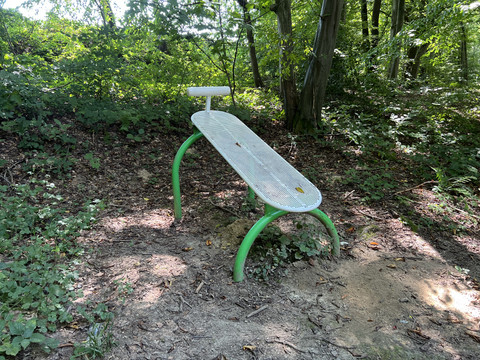 Trimm-Dich-Pfad Grüne Parkbank im Wald, umgeben von Bäumen und Laub auf dem Boden, lädt zur Rast in der Natur ein.