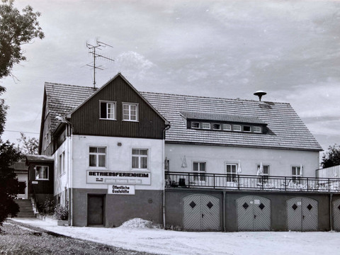 Černobílá fotografie dvoupatrové budovy s nápisy "Betriebsferienheim" a "Öffentliche Gaststätte", obklopené stromy.