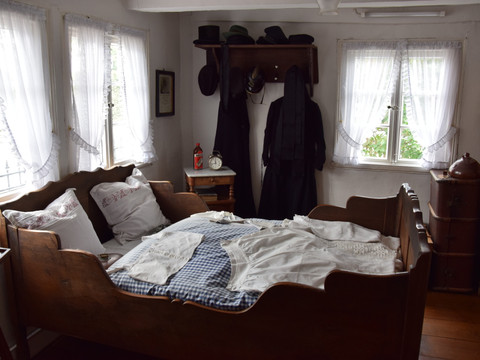 Bauernhofmuseum <p>Historisches Schlafzimmer mit antikem Holzbett und traditioneller Kleidung vor weißen Vorhängen.</p>