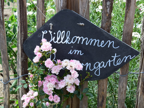 Bauernhofmuseum Willkommensschild mit Rosenblüten vor einem rustikalen Holzzaun im Garten.