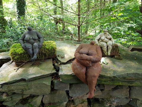 Galerie Sattelgut Drei Skulpturen sitzende Figuren aus Ton auf einer steinernen Mauer in einem bewaldeten Gebiet.