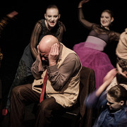 Theater der Keller Sechs in fantasievollen Kostümen gekleidete Schauspielende, maskiert, umkreisen einen Mann, der die Ohren zuhaltend auf einem Stuhl sitzt. Ihre ausdrucksstarken Gesten lassen eine intensive dramatische Szene vermuten.Six masked actors dressed in imaginative costumes circle a man sitting in a chair with his ears covered. Their expressive gestures suggest an intense dramatic scene.