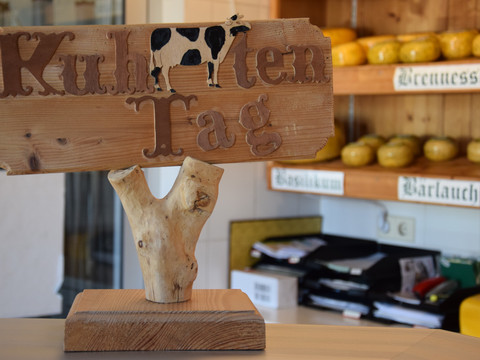 Thomashof Burscheid <p>Holzschild mit Text "Kuhlturen Tag" und Kuh, im Hintergrund Käselaibe auf Regalen.</p>