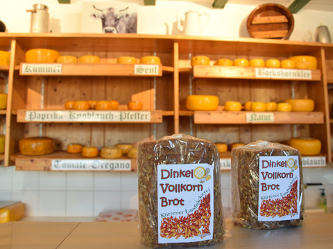 Thomashof Burscheid  Zwei Dinkelvollkornbrote vor einem Regal voller Käselaibe in einem rustikalen Geschäft.