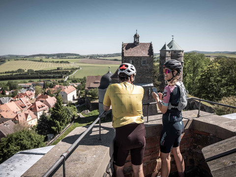 View from Stolpen Castle Zwei Radfahrer mit Helmen stehen auf einer Mauer der Burg Stolpen und blicken auf eine malerische Landschaft mit Feldern und einem Dorf mit roten Dächern.Two cyclists with helmets stand on a wall of Stolpen Castle and look out over a picturesque landscape of fields and a village with red roofs.Dva cyklisté s helmami stojí na zdi hradu Stolpen a dívají se na malebnou krajinu s poli a vesnicí s červenými střechami.Dwóch rowerzystów w kaskach stoi na ścianie zamku Stolpen i patrzy na malowniczy krajobraz z polami i wioską z czerwonymi dachami.Twee fietsers met helmen staan op een muur van Kasteel Stolpen en kijken uit over een schilderachtig landschap met velden en een dorp met rode daken.Due ciclisti con il casco sono in piedi su una parete di Castel Stolpen e guardano un paesaggio pittoresco con campi e un villaggio dai tetti rossi.