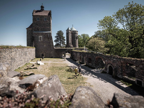 The fourth courtyard at Stolpen Castle Historische Burg Stolpen mit zwei Türmen aus Stein, umgeben von grünen Bäumen; zwei Personen spazieren im Innenhof bei sonnigem Wetter.Historic Stolpen Castle with two stone towers, surrounded by green trees; two people strolling in the courtyard in sunny weather.Historický hrad Stolpen se dvěma kamennými věžemi, obklopený zelenými stromy; dva lidé se za slunečného počasí procházejí po nádvoří.Zabytkowy zamek Stolpen z dwiema kamiennymi wieżami, otoczony zielenią drzew; dwoje ludzi spacerujących po dziedzińcu przy słonecznej pogodzie.Historisch Kasteel Stolpen met twee stenen torens, omringd door groene bomen; twee mensen wandelen op de binnenplaats bij zonnig weer.Storico castello di Stolpen con due torri in pietra, circondato da alberi verdi; due persone passeggiano nel cortile con il sole.