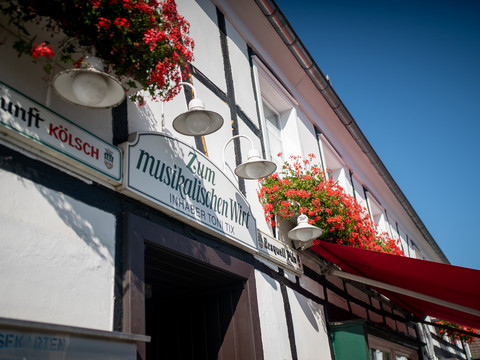 Außenansicht Fachwerkhaus mit Schildern, roten Blumen und Sonnendach; traditionell und einladend gestaltet.
