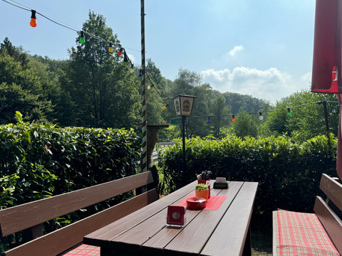 Landhaus Orbach Biergarten Sonniger Biergarten mit Holzbänken und -tischen, umgeben von grünen Büschen und bunten Lichterketten.
