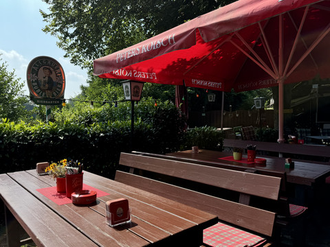 Landhaus Orbach Biergarten Ein idyllischer Biergarten mit roten Tischen und Sonnenschirmen, umgeben von üppigem Grün.
