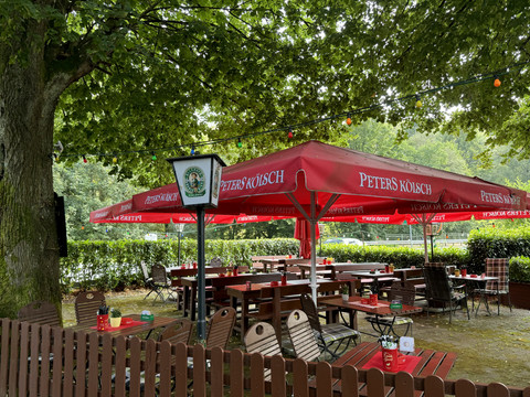Landhaus Orbach Biergarten Gemütlicher Biergarten unter grünen Bäumen, mit roten Sonnenschirmen und Holztischen, einladend gedeckt.