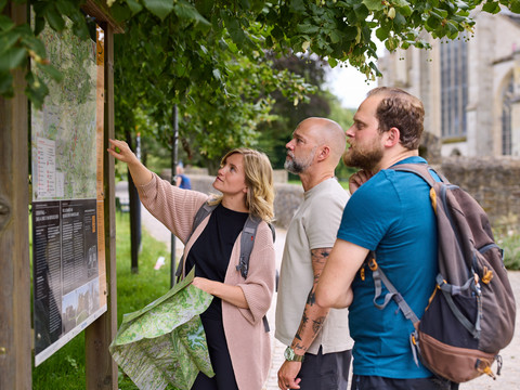 Bergischer Weg in Altenberg <p>Eine Gruppe betrachtet interessiert eine Informationstafel im Freien, umgeben von Bäumen.</p>