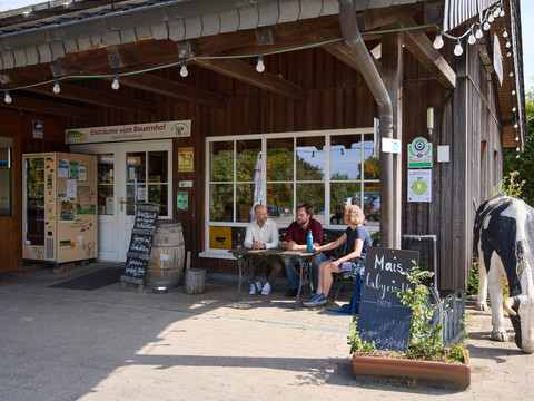 Hofladen Hielscher Hof <p>Personen sitzen an einem Tisch vor einem rustikalen Bauernhofladen; dekoriert mit Schildern und Pflanzen.</p>