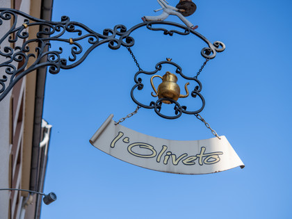 L'Oliveto Schild Schild "l'Oliveto" an verschnörkelter Aufhängung vor blauem Himmel