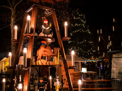 Zusehen ist im Vordergrund eine beleuchtete Holzpyramide. Im Hintergrund ist ein weihnachtlicher, bedachter Stand.
