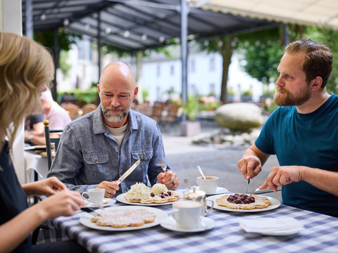 Bergische Waffeln Ein Trio genießt auf einer Terrasse unter einer Überdachung frische Waffeln mit Sahne und Kirschen.