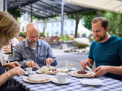 Bergische Waffeln Drei Personen genießen Kaffee und Waffeln an einem Gartentisch eines Cafés mit entspannter Atmosphäre.