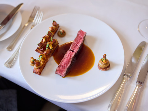 Restaurant Altenberger Hof  Roastbeef mit Jus, garniert mit Kartoffelwürfeln und Pilzen auf weißem Teller, Besteck rechts.