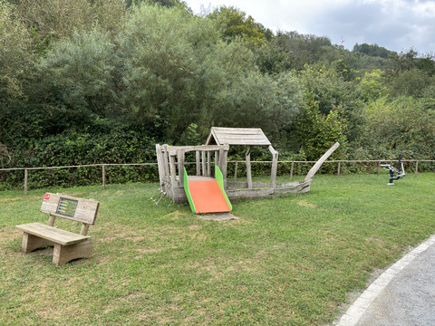 Spielplatz am Aggerstrand Ründeroth Holzspielplatz in Form eines Schiffs mit Rutsche, umgeben von grüner Natur und mit einer Parkbank.