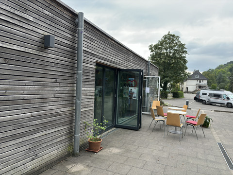 Aggerstrand in Ründeroth <p>Moderner Holzbau mit großen Glasfenstern, davor bunte Stühle und Tisch auf gepflasterter Terrasse.</p>
