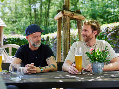 Holsteins Mühle  Zwei Männer sitzen lächelnd an einem Holztisch im Freien mit Bier und einer Blumendekoration darauf.