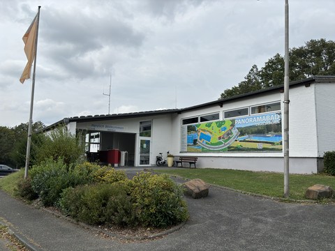 Panoramabad Panoramabad Engelskirchen: Eingangsbereich mit Willkommensbanner, Werbung und Gehweg, trüb bewölkter Himmel.