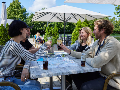 Pause im Biergarten  <p>Vier Personen stoßen mit Getränken an und lächeln im Freien an einem Tisch mit einer Karte.</p>