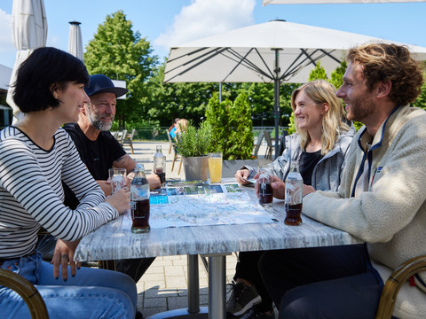 Pause im Biergarten  Eine Gruppe von vier Personen sitzt fröhlich an einem Tisch im Freien und genießt Erfrischungsgetränke.