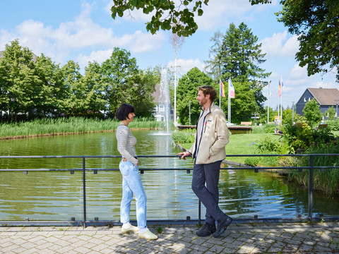 Pause im Biergarten  Zwei Personen unterhalten sich vor einem idyllischen Teich mit Wasserfontäne, umgeben von Bäumen.