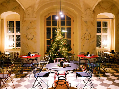 Gemütlicher Raum mit weihnachtlich geschmücktem Baum, runden Tischen und bunten Stühlen auf einem Schachbrettboden, warm beleuchtet.