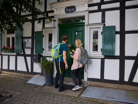 Restaurant Zur Post Zwei Wanderer mit Rucksäcken stehen vor einem Fachwerkhaus und öffnen eine grüne Haustür.