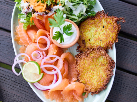 Biergarten Rusticus Lachstatar mit Zwiebeln, Kartoffelpuffer, Salatgarnitur und Dipsauce auf einem weißen Teller.