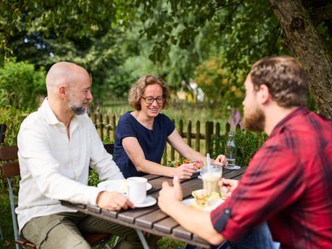 Biergarten Rusticus Eine Gruppe von drei Personen sitzt an einem Tisch draußen, sie genießen Getränke und sind in ein Gespräch vertieft.