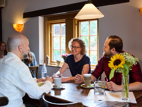 Restaurant Rusticus Gemütliches Café, drei Personen unterhalten sich bei Kaffee, Holztisch und Sonnenblumen im Vordergrund.