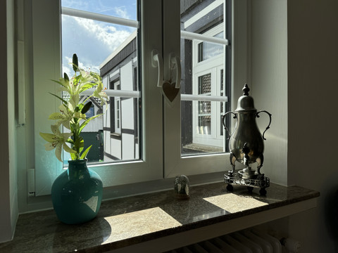 Ansicht Fensterbank mit Dröppelminna Ein Fensterbrett mit einer blauen Vase und Blumen, einer stilvollen Teekanne und einem sonnigen Ausblick.