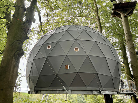 Baumhaus im Hochseilgarten K1 Eine geodätische Kuppel in einem Baum umgeben von grünen Blättern mit runden Fenstern in der Struktur.