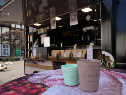 Sheka Forest Kaffee Recup Ein brauner und ein grüner Pfandbecher der Marke Recup stehen vor dem Kaffeemobil auf einem Tisch