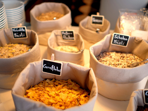 Frühstück (1).jpg Verschiedene Müslisorten und Cornflakes in Stoffbehältern, beschriftet mit kleinen Tafeln auf einem Tisch.